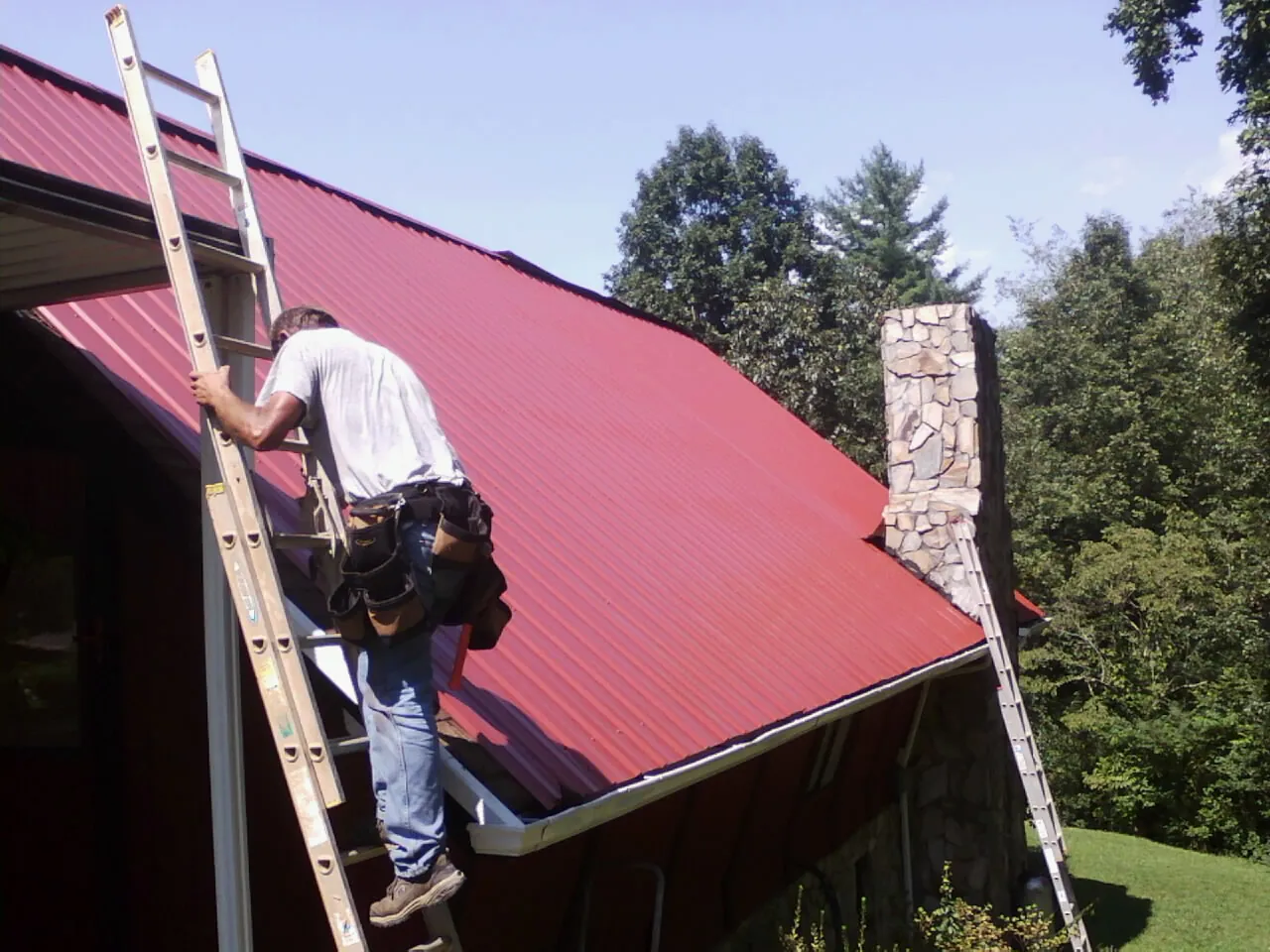 Roofer on a ladder beside a red metal roof and stone chimney — Soffit & Fascia Repair by Roofing Brothers in Frenchglen